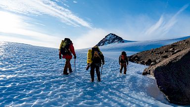 Expeditioners walk through a dramatic, mountainous landscape. Rocky mountains rise out of an icy plain.