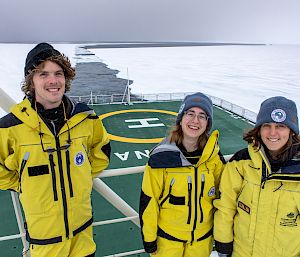 3 expeditioners on the deck of a ship. Behind them is the path through the ice that has been created by the icebreaker ship.