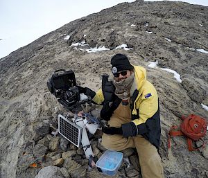 Expeditioner sitting on rocks next to electrical equipment and remote camera.