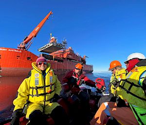 Expeditioners wearing lifejackets sit in an inflatable boat. A large red ship is behind, lifting cargo with a crane.