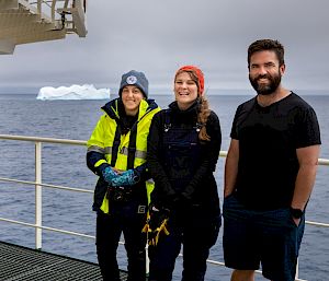 4 expeditioners pose for a photo on the deck of a ship. There is an iceberg in the distance behind them.