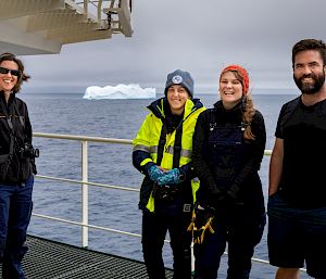 4 expeditioners pose for a photo on the deck of a ship. There is an iceberg in the distance behind them.