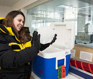 A scientist in black and yellow cold weather gear and gloves holds a piece of ice above a blue esky.