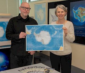 2 people hold a map of Antarctica. Several other maps are on the walls around them.