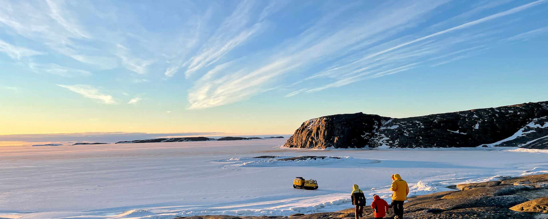 Three expeditioners walk down some rocks towards a large expanse of sea ice. A yellow Hagglund vehicle can be seen parked out on the ice.