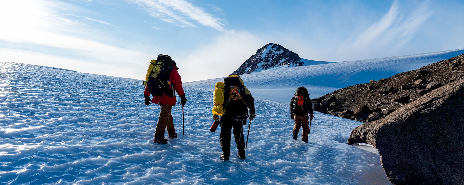 Expeditioners walk through a dramatic, mountainous landscape. Rocky mountains rise out of an icy plain.