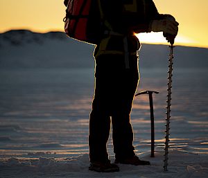 An expeditioner stands in the light of the rising sun with a long drill.
