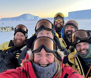 A group of expeditioners pose with penguins in the distance.