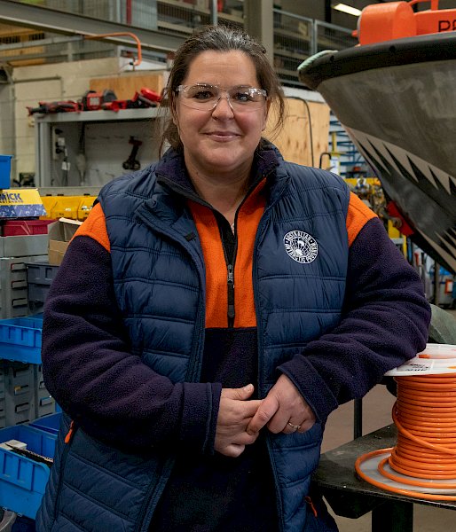 A woman in high vis and a puffer vest smiles in an electrical workshop