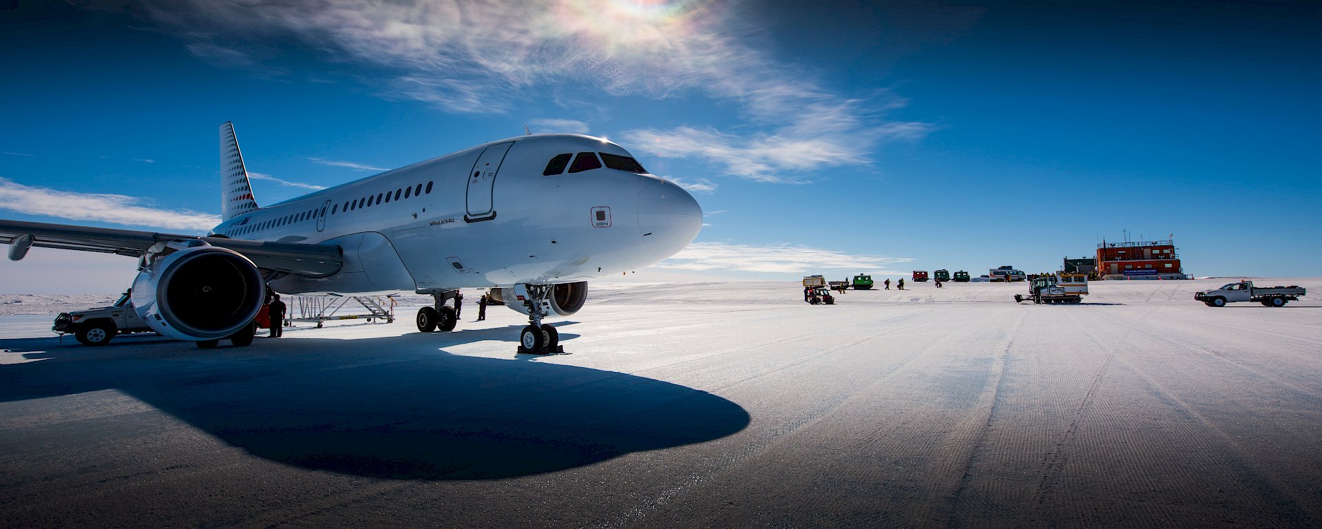 Airbus A319 parked on runway with aerodrome operations building and vehicles in distance behind and large sun overhead.