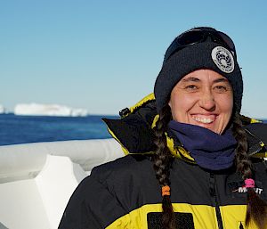 An expeditioner stands on a boat and smiles at the camera. In the background is the blue sky and ocean with an iceberg.
