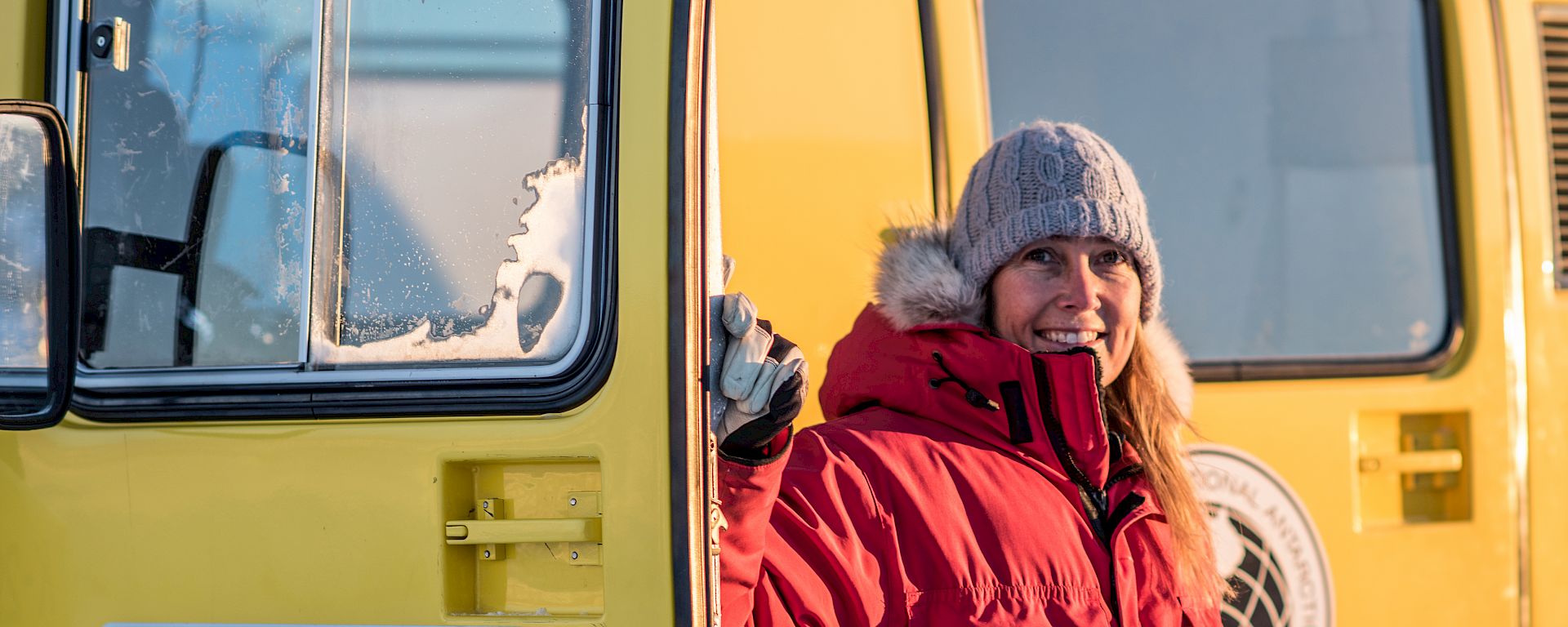 An expeditioner in red coat standing beside a yellow over-snow vehicle.