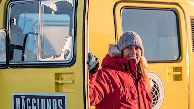 An expeditioner in red coat standing beside a yellow over-snow vehicle.