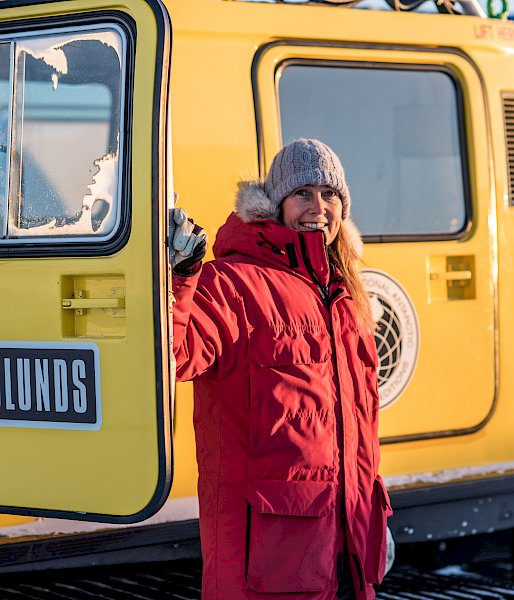 An expeditioner in red coat standing beside a yellow over-snow vehicle.