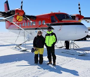 2 expeditioners stand on the sea ice in front of a small red and white aircraft.