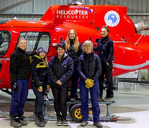 A group of smiling expeditioners pose in front of a helicopter.