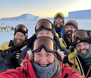 A group of expeditioners pose for a selfie with penguins in the distance.