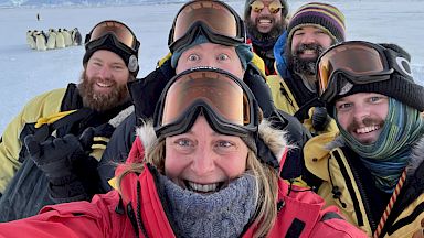 A group of expeditioners pose for a selfie with penguins in the distance.