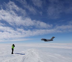 A large aircraft takes off from an ice runway under a blue sky. A person in hi-vis stands in the foreground.