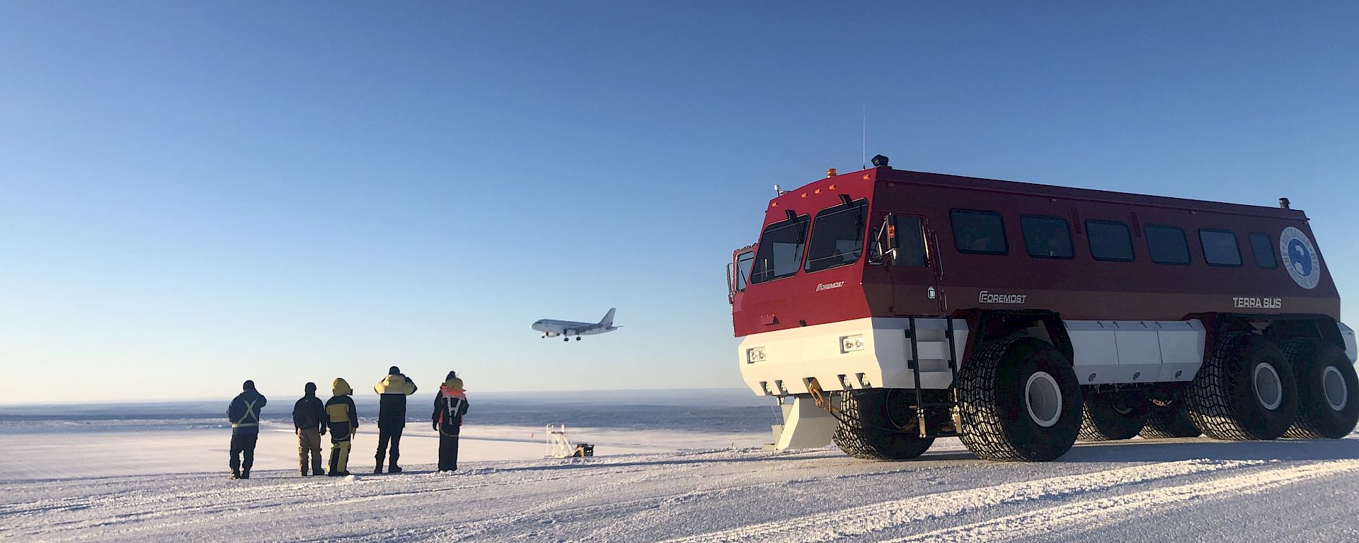 A group of people standing next to a bus, watching a plane land on the ice.