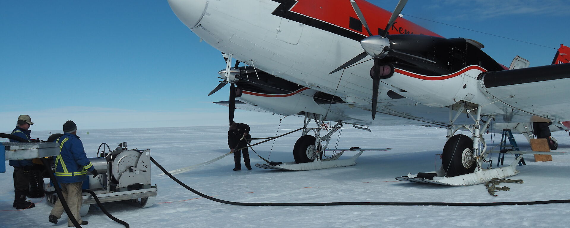 Top right of picture, red and white plane (forward of wings). On white snow runway. Blue sky above. Two men standing at fuel pump to left of picture with hose running towards the plane