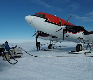 Top right of picture, red and white plane (forward of wings). On white snow runway. Blue sky above. Two men standing at fuel pump to left of picture with hose running towards the plane