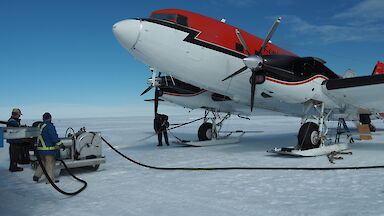 Top right of picture, red and white plane (forward of wings). On white snow runway. Blue sky above. Two men standing at fuel pump to left of picture with hose running towards the plane