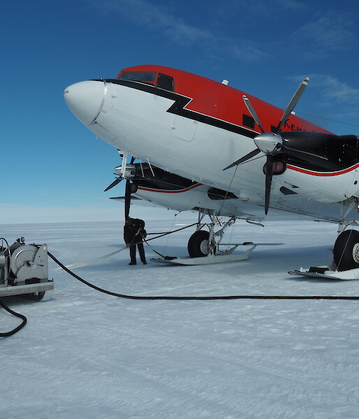 Top right of picture, red and white plane (forward of wings). On white snow runway. Blue sky above. Two men standing at fuel pump to left of picture with hose running towards the plane