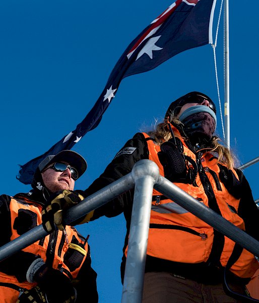Low angle of expeditioners wearing high vis, on deck of building with Australian flag behind