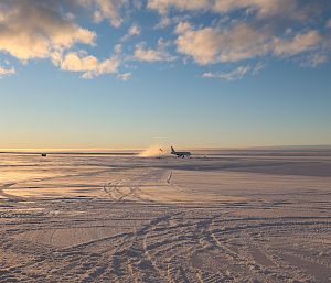 A plane lands on a wide, flat plain of snow and ice. A cloud of snow is kicked up in its wake.