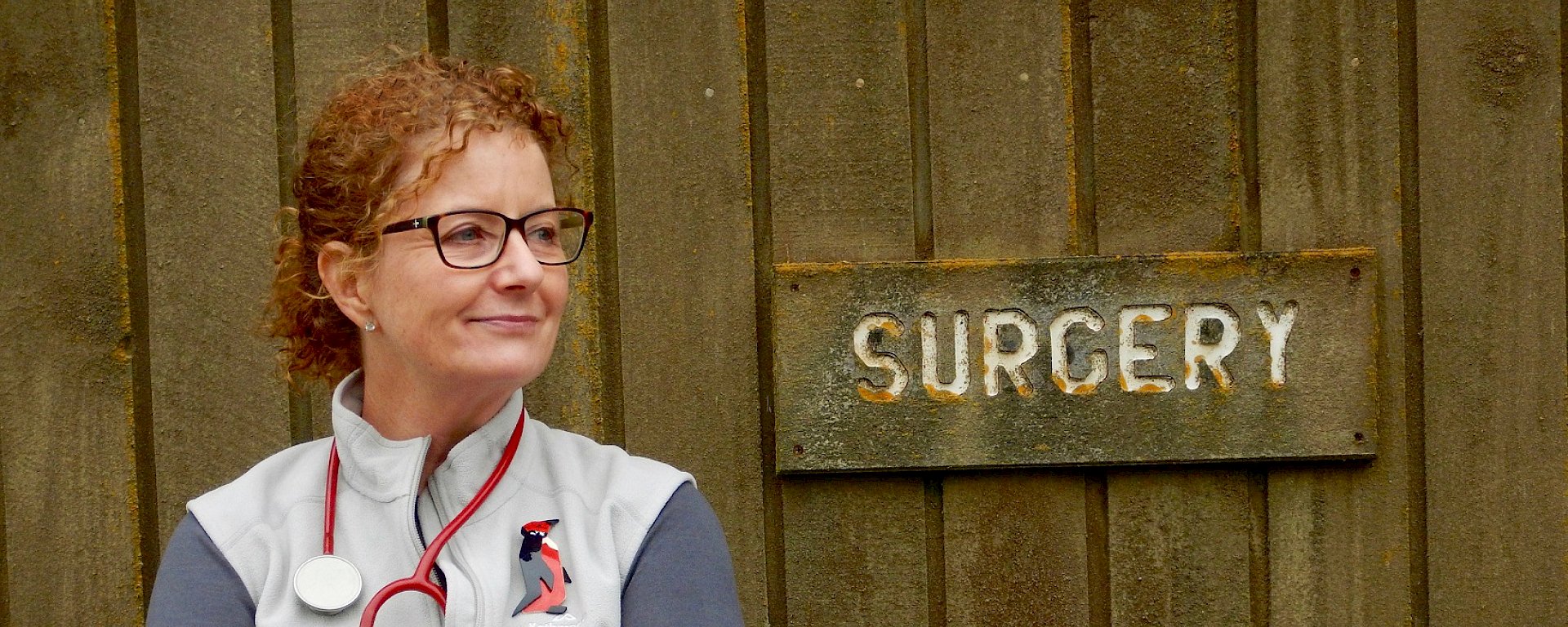 Station doctor with stethoscope around her neck standing beside lichen-encrusted sign that reads ‘surgery’.