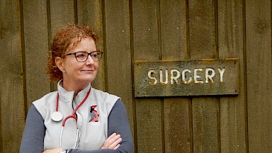 Station doctor with stethoscope around her neck standing beside lichen-encrusted sign that reads ‘surgery’.
