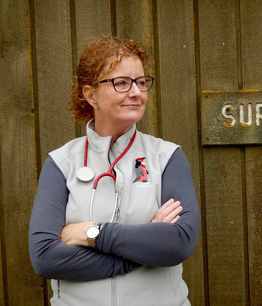 Station doctor with stethoscope around her neck standing beside lichen-encrusted sign that reads ‘surgery’.