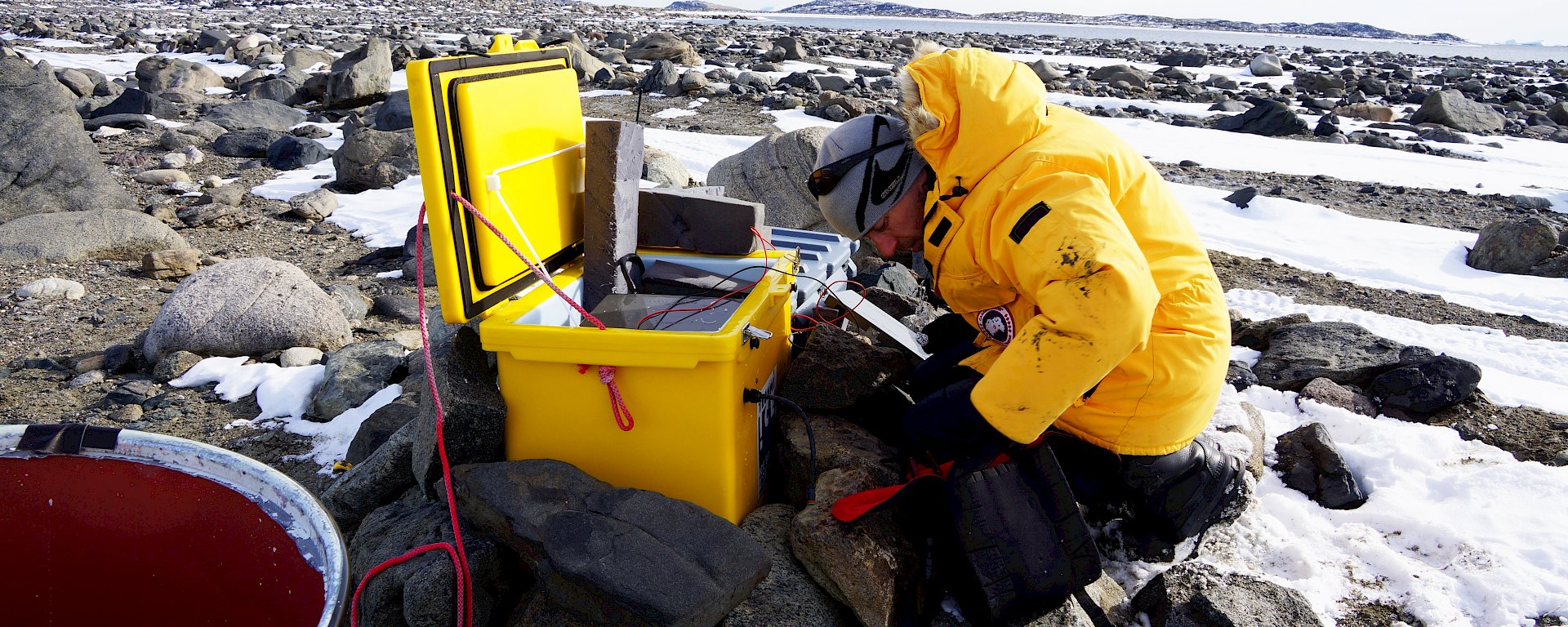 Man kneeling in front of yellow plastic box adjusting electronics equipment, on snow and rock covered ground.