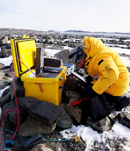 Man kneeling in front of yellow plastic box adjusting electronics equipment, on snow and rock covered ground.