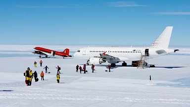 Large white aircraft and smaller red aircraft on the ice with disembarked passengers milling about.