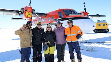 A group of five smiling people stand in front of a red and white plane on the sea ice.