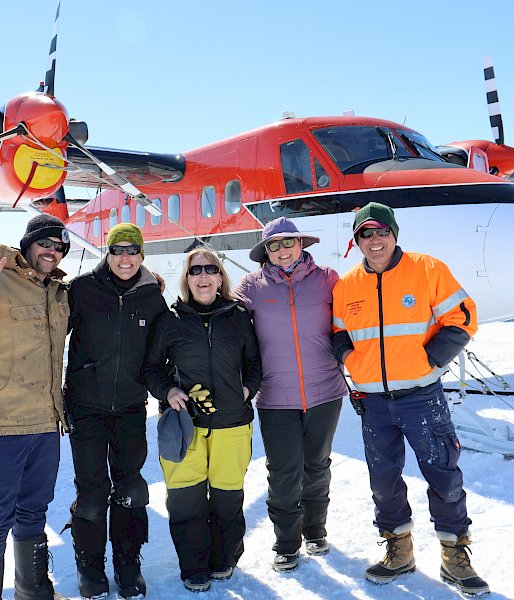 A group of five smiling people stand in front of a red and white plane on the sea ice.