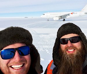 Two bearded men posing with plane on runway.