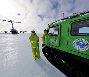 A large aircraft sits on the ice under a blue sky. 2 expeditioners stand next to a green vehicle in the foreground.