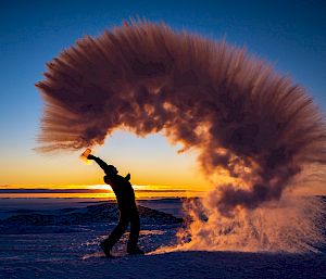 An expeditioner stands on the ice with the setting sun behind. They throw water into the air which is captured by the photo as it vaporises and freezes in the cold.