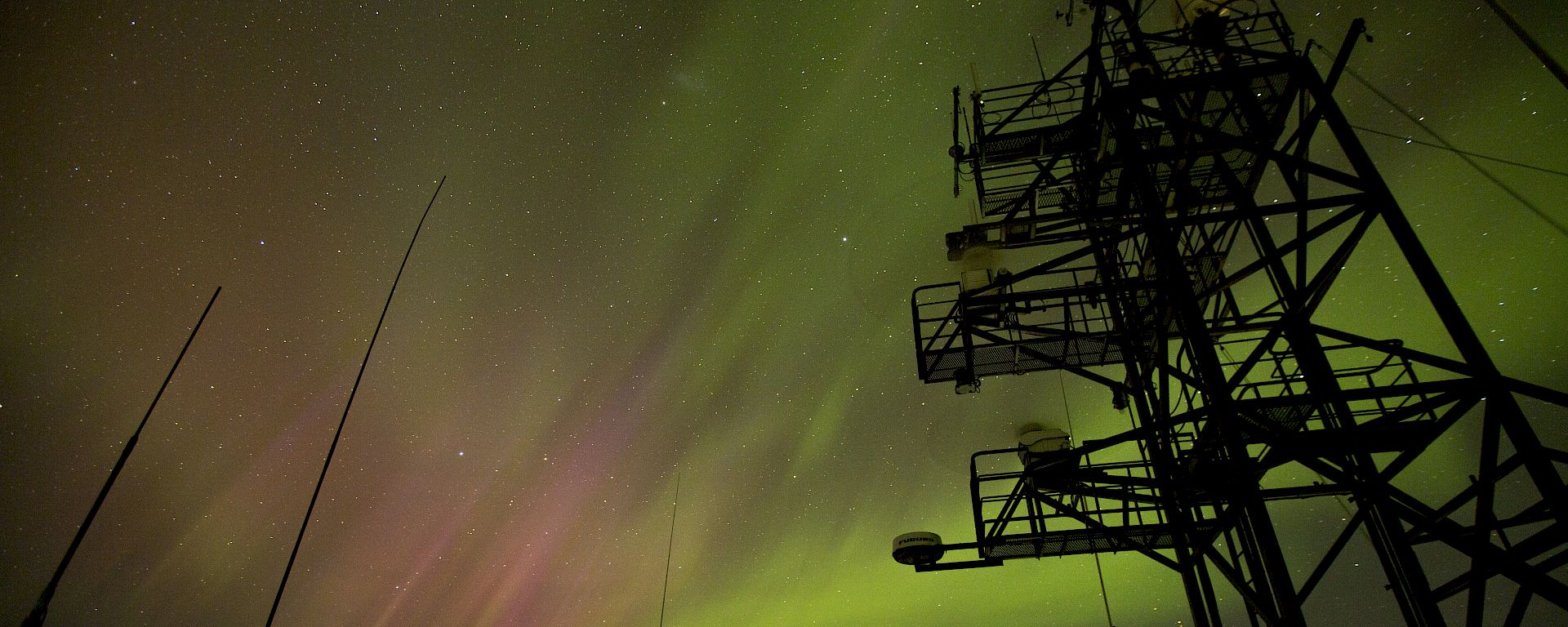 Green and pink aurora viewed from ship with silhouettes of antennae and masts.