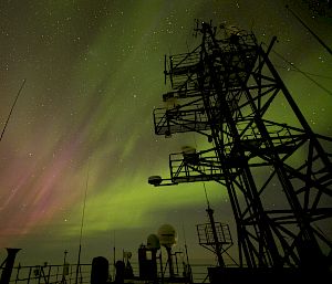 Green and pink aurora viewed from ship with silhouettes of antennae and masts.