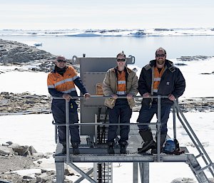 Three people standing on metal structure with ice and ocean in background.