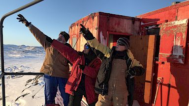 Three tradespeople stand outside a red field hut.