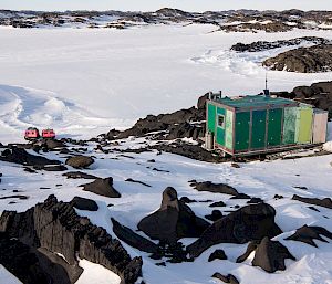 A small, green hut in a rocky, snow-covered landscape.