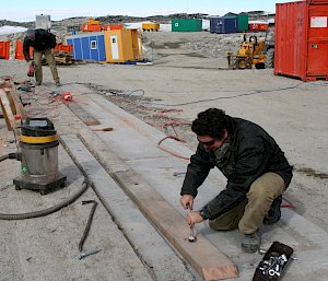 Two men working on piece of wood on concrete slab outdoors.