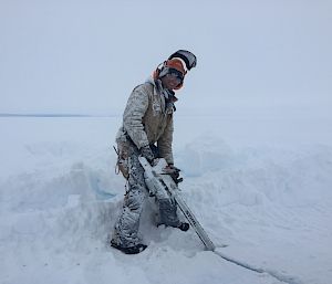 An expeditioner with snow-covered clothing holds a chainsaw pointing down into the ice.