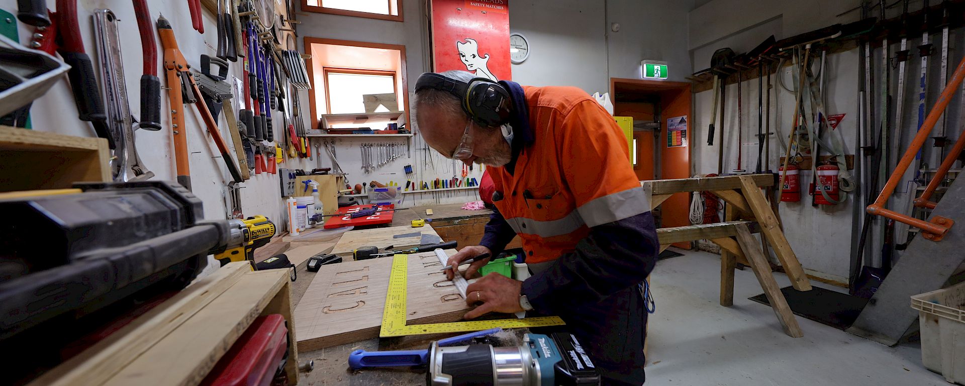 An expeditioner in hi-vis works on a wooden sign in a workshop.