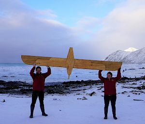Two people on a snowy beach hold up a wooden albatross.